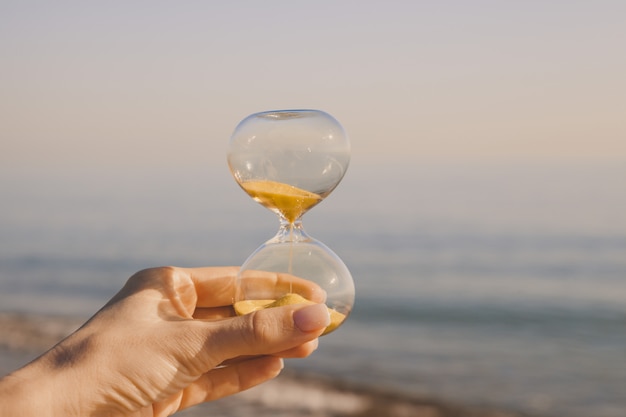 Weibliche Hand mit einer Sanduhr gegen den Sandstrand eines heißen Landes, Zeit läuft durch Finger, Konzept zurück in die Vergangenheit