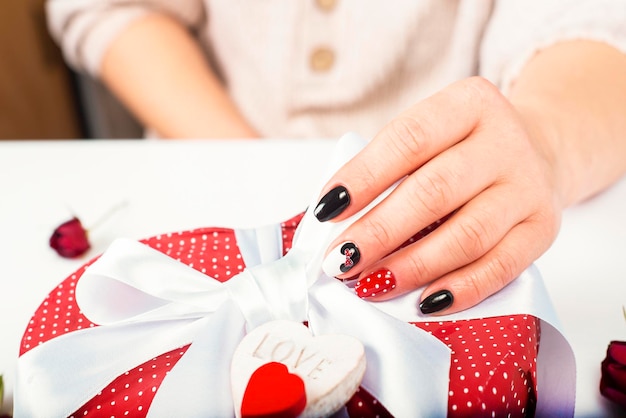 Weibliche Hand mit Maniküre-Gel-Lak löst eine Schleife auf einer Geschenkkiste mit Herzen Closeup-Geschenk für