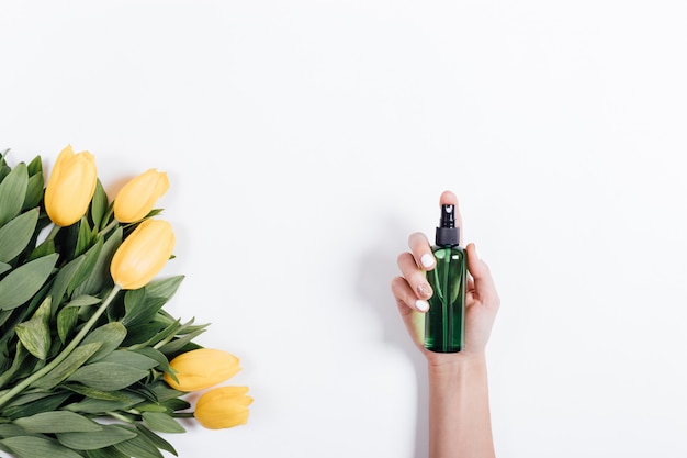 Weibliche Hand mit Maniküre hält eine grüne Plastikflasche Wasser nahe gelben Tulpen, Ansicht von der Spitze