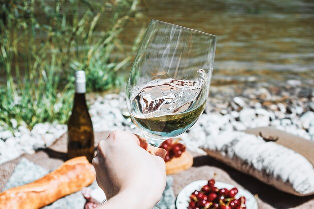 weibliche Hand mit Weißweinglas an der Flussküste und Picknickhintergrund