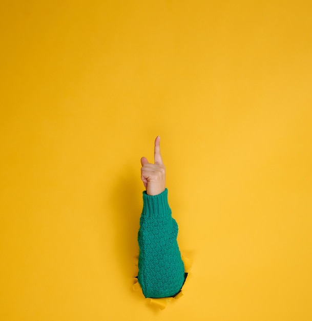 Foto weibliche hand ragt aus einem zerrissenen loch in einem gelben papierhintergrundteil des körpers zeigt mit dem zeigefinger nach oben kopierraum