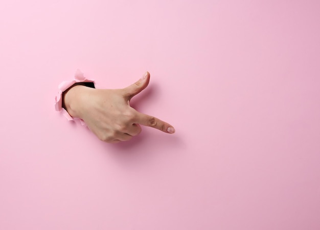 Weibliche Hand ragt aus einem zerrissenen Loch in einem rosa Papierhintergrund, ein Teil des Körpers zeigt mit dem Zeigefinger zur Seite