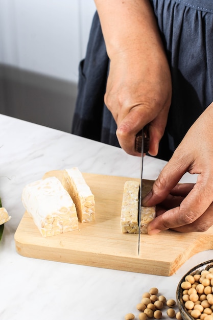 Weibliche Hand Schneiden von Tempeh oder Tempe auf Schneidebrett aus Holz mit Messer. Tempe ist ein fermentiertes Sojaprodukt, das ursprünglich aus Indonesien stammt. Kochprozess Herstellung von Stir Fry Tempeh (Orek)