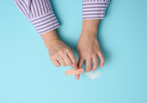 Weibliche Hand und braunes medizinisches Pflaster zur Behandlung von Verletzungen und Schnitten auf der Haut, blauer Hintergrund