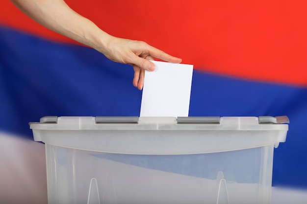 Weibliche Hand wirft Stimmzettel in die Wahlurne. Russische Flagge im Hintergrund.