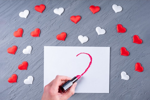 Weibliche Hand zeichnet mit Lippenstift eine halbe Herzform auf weißem Papier auf einem grauen hölzernen Hintergrund