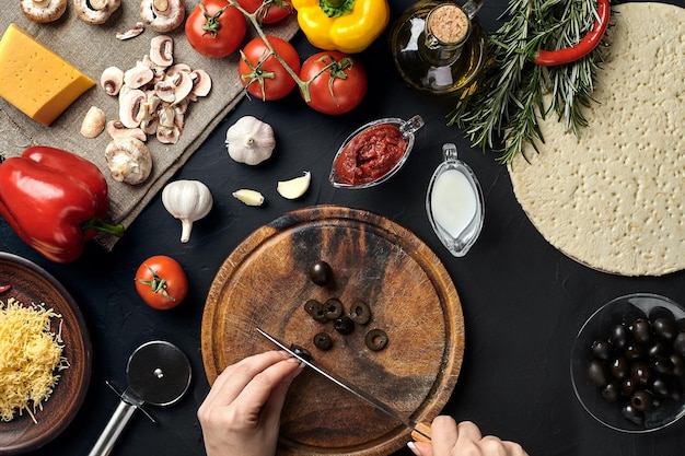 Weibliche handgeschnittene Oliven auf Holzbrett auf Küchentisch um Lügenzutaten für Pizzagemüsekäse und Gewürze Beschneidungspfad eingeschlossen