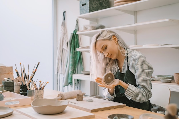 Weibliche Handwerkerin mit Schürze arbeitet an Keramikgeschirr mit Handwerkzeugen in einer Töpferwerkstatt Junge Frau, die Lehmprodukte in einer Keramikwerkstatt herstellt Lächelndes Mädchen im Kunststudio