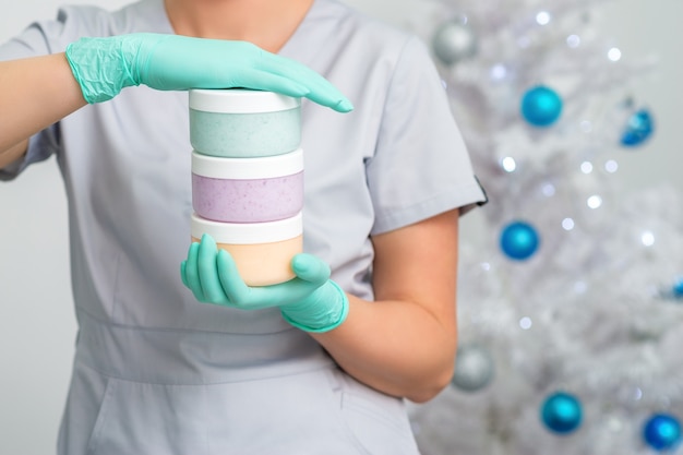 Weibliche Kosmetikerin, die drei leere Gläser mit Zuckerpaste auf Weihnachtsbaumhintergrund hält
