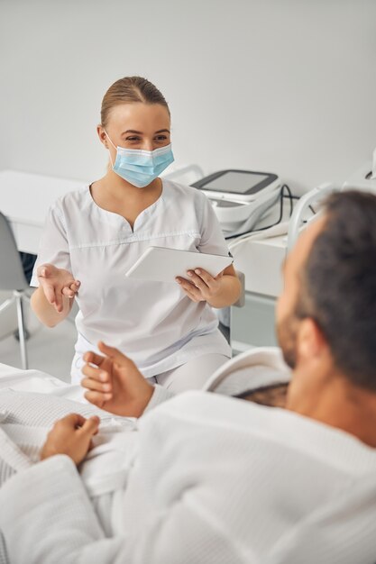 Weibliche Kosmetikerin in medizinischer Gesichtsmaske, die einen elektronischen Pad-PC hält, während sie mit einem männlichen Patienten spricht