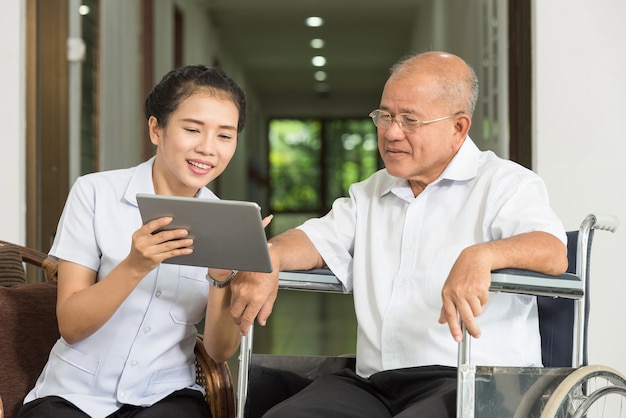 Weibliche Krankenschwester, die über digitaler Tablette mit älterem Mann im Rollstuhl am Pflegeheim sich bespricht