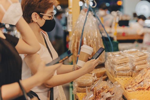 Weibliche Kunden in Schutzmaske machen kontaktloses mobiles Bezahlen im Einzelhandelsgeschäft.