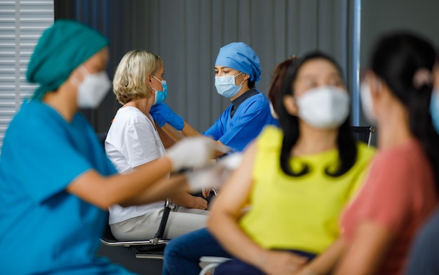 Weibliche Oktorin trägt Gesichtsmaskengummihandschuhe und blaue Krankenhausuniform, die der kaukasischen Frau einen Impfstoff injiziert, während andere Krankenschwestern weibliche Patienten in der Warteschlange zusammen in der Klinik impfen, verschwommener Vordergrund.