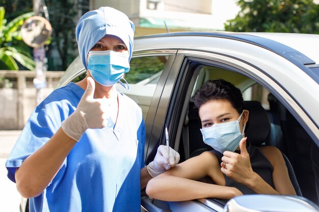 Weibliche Patientin sitzt im Auto und kaukasische Ärztin trägt Gesichtsmaske halten Kovid 19 Spritzennadel-Impfstoffständer halten Daumen zusammen Blick auf die Kamera in Fahrt durch die Impfschlange