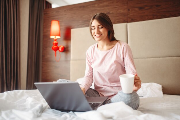 Weibliche Person trinkt Kaffee und benutzt Laptop im Bett