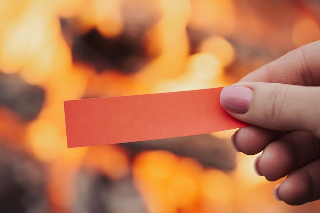 Weibliche rechte Hand, die ein rotes kleines Blatt Papier mit Kopierbereich hält Nahaufnahme eines leeren Lagerfeuers im Hintergrund