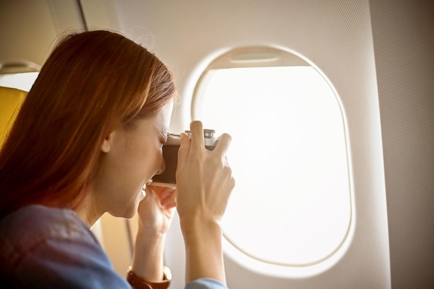 Weibliche Reisende, die eine Retro-Kamera verwenden, um ein Himmelsfoto durch das Fenster des Flugzeugs zu machen