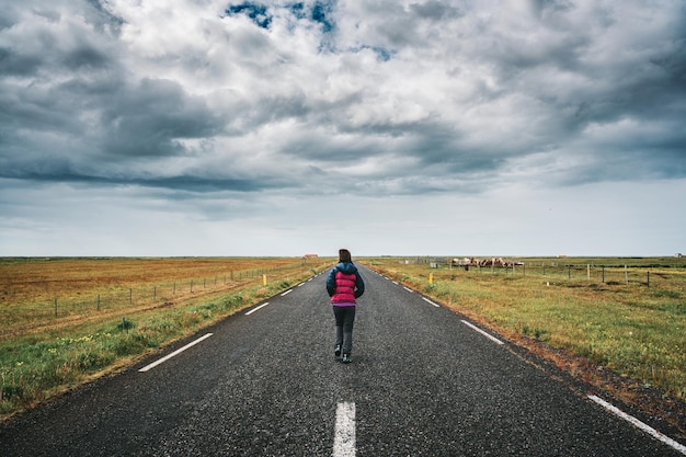 Weibliche Touristen, die im Sommer auf einer geraden Straße durch Ackerland und stürmischen Himmel auf dem Land laufen