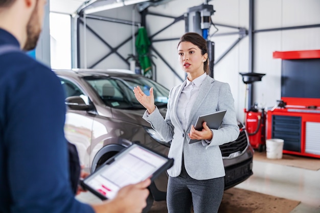 Weiblicher Autoverkäufer, der in der Garage des Autosalons steht und mit einem Mechaniker über das Reparieren des Autos spricht