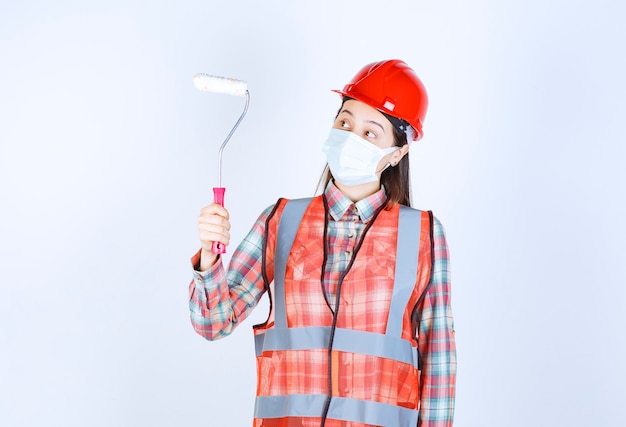 Weiblicher Bauingenieur in Sicherheitsmaske und rotem Helm, der eine Trimmrolle zum Malen hält und verwirrt sieht.