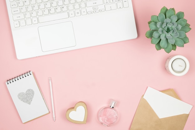 Weiblicher Bürodesktop mit Bürozubehör auf rosa Oberfläche. Damenarbeitsbereich mit Sukkulenten, Kerzen und Kosmetika.