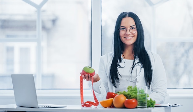 Weiblicher Ernährungswissenschaftler im weißen Mantel, der Apfel mit messendem Band hält.