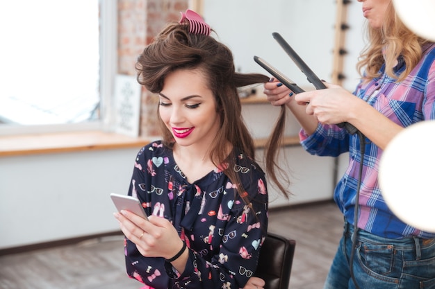 Weiblicher Friseur, der Haarglätter für langes Haar einer lächelnden jungen Frau mit Smartphone in der Umkleidekabine anwendet