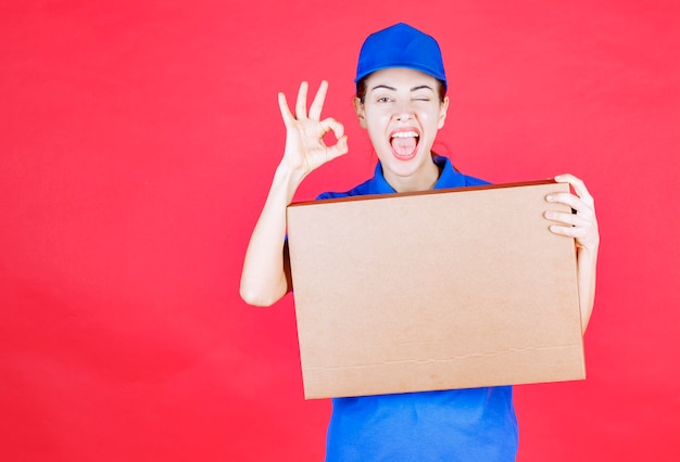 Weiblicher Kurier in blauer Uniform, der einen Pizzakarton zum Mitnehmen aus Pappe hält und ein Genusszeichen zeigt.