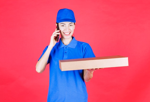 Weiblicher Kurier in blauer Uniform, der einen Pizzakarton zum Mitnehmen aus Pappe hält und mit dem Telefon spricht.