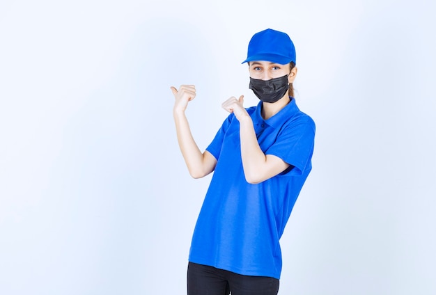 Weiblicher Kurier in Maske und blauer Uniform, die etwas hinter sich zeigen.