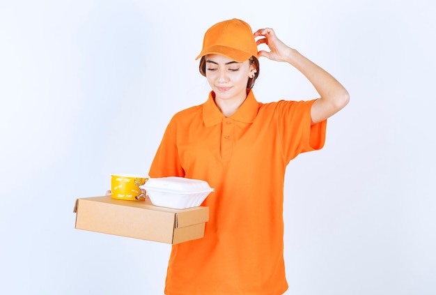 Weiblicher Kurier in orangefarbener Uniform, der gelbe und weiße Takeaway-Boxen mit einem Papppaket hält und verwirrt und nachdenklich aussieht