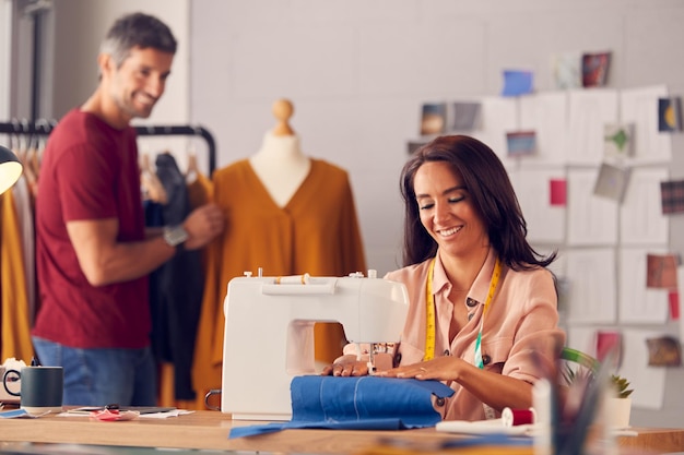 Weiblicher Modedesigner arbeitet an Nähmaschine als männlicher Kollege arbeitet im Hintergrund