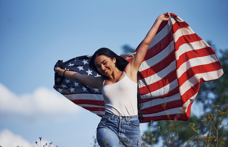Foto weiblicher patriot läuft mit usa-flagge in den händen draußen im feld gegen blauen himmel.