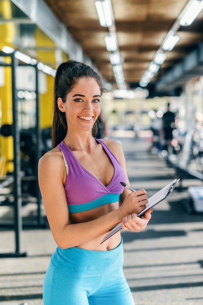 Weiblicher Personal Trainer mit Pferdeschwanz und gekleidet in Sportbekleidung, die auf Zwischenablage schreibt Ergebnisse des Trainings beim Stehen in einem Fitnessstudio und beim Betrachten der Kamera.