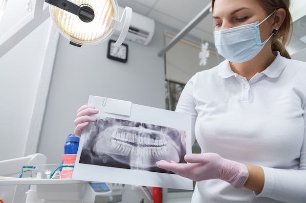 Weiblicher Zahnarzt, der medizinische Gesichtsmaske trägt, die zahnärztliche Röntgenaufnahme zur Kamera zeigt