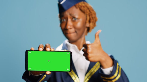 Weibliches Flugzeugbesatzungsmitglied, das auf die Greenscreen-Anzeige über blauem Hintergrund vor der Kamera zeigt. Junge Stewardess mit isoliertem Copyspace und Chroma-Key-Vorlage in Luftfahrtuniform