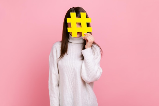Weibliches Gesicht mit Social-Media-Hashtag-Symbol, Empfehlung, trendigen Inhalten zu folgen, Blog, mit weißem Pullover im lässigen Stil. Studioaufnahme im Innenbereich isoliert auf rosa Hintergrund.