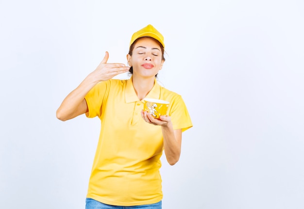 Weibliches Mädchen in gelber Uniform, das eine gelbe Nudelschale zum Mitnehmen hält und das Produkt riecht.