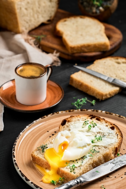 Weich gekochtes (pochiertes) Ei auf einer mit Buttercreme und Kräutern bedeckten Brotscheibe auf einer Tonplatte auf einem schwarzen Holztisch. Espresso und Laib geschnittenes Brot an der verschwommenen Wand. Frühstücksidee