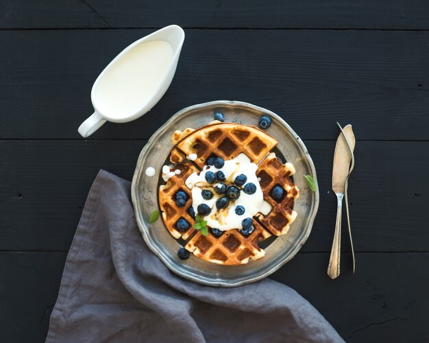 Weiche belgische Waffeln mit Blaubeeren, Honig und Schlagsahne auf Weinlesemetallplatte über schwarzem Holztisch, Draufsicht.