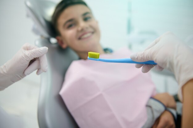 Weicher Fokus auf eine Zahnbürste in der Hand eines Arztes Zahnarzt lehrt einen unscharfen lächelnden gutaussehenden Jungen, der im Zahnarztstuhl über Mundpflege und tägliche Zahnhygiene sitzt. Medizinisches Konzept