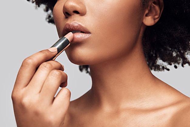 Weicher Lippenstift. Nahaufnahme einer jungen Afrikanerin, die Lippenstift aufträgt, während sie vor grauem Hintergrund steht