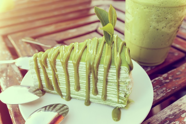 Weiches Teeblatt im Vintage-Stil, gestickt auf Matcha-Kuchen in weißer Platte, in der Nähe eines Glases eisgrünen Tees auf einem alten Holztisch in der Abendsonne