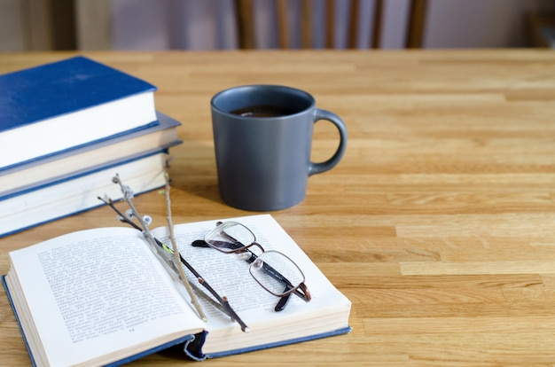 Weidenzweige und Gläser liegen auf einem offenen Buch. Auf einem Holztisch liegt ein Stapel Bücher und eine Tasse Kaffee.