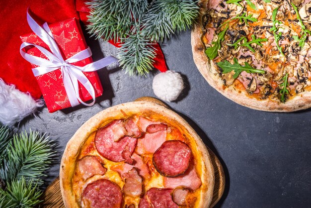 Weihnachten dekorierter Hintergrund mit Peperoni- und Pilzpizza, Lieferung und Restaurant Xmas Lunch- und Partymenü, Santa liefert Pizza zu Weihnachten, schwarzer Betonhintergrund Draufsicht Kopienraum