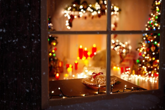 Weihnachten dekoriertes Zimmer Interieur mit Girlandenlichtern, Kerzen und Tannenbaum. Winter-mattierter Fensterrahmen. Weihnachtsfeiertags-Lebkuchenkekse für den Weihnachtsmann am Tisch