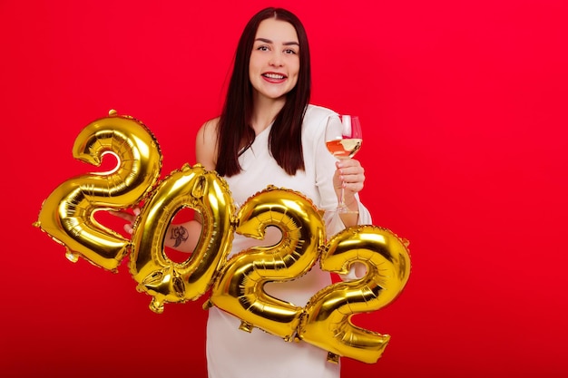 Weihnachten, Feiertag - eine junge lächelnde Frau in einem weißen Kleid hält die Zahlen 2022 und ein Glas Wein