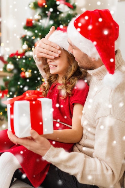 weihnachten, feiertage, familien- und menschenkonzept - lächelnder vater und tochter in weihnachtsmützen, die geschenkbox halten und zu hause die augen bedecken