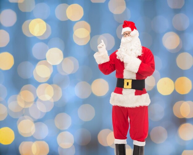 weihnachten, feiertage, gesten und menschenkonzept - mann im weihnachtsmannkostüm, der mit dem finger auf den hintergrund der blauen lichter zeigt