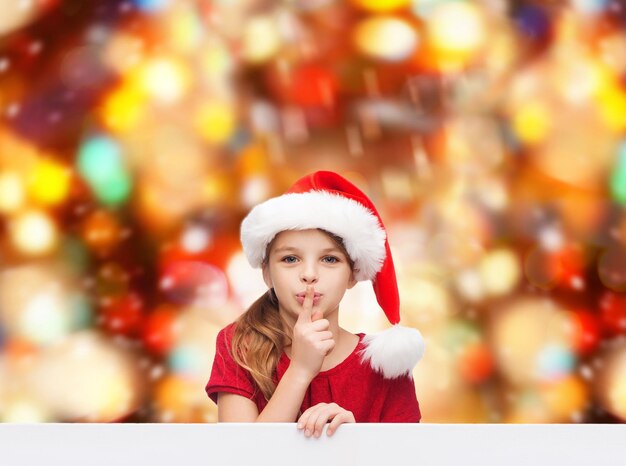 weihnachten, feiertage, kindheit und menschenkonzept - lächelndes kleines mädchen in weihnachtsmannmütze mit dem finger auf den lippen über rotem lichthintergrund